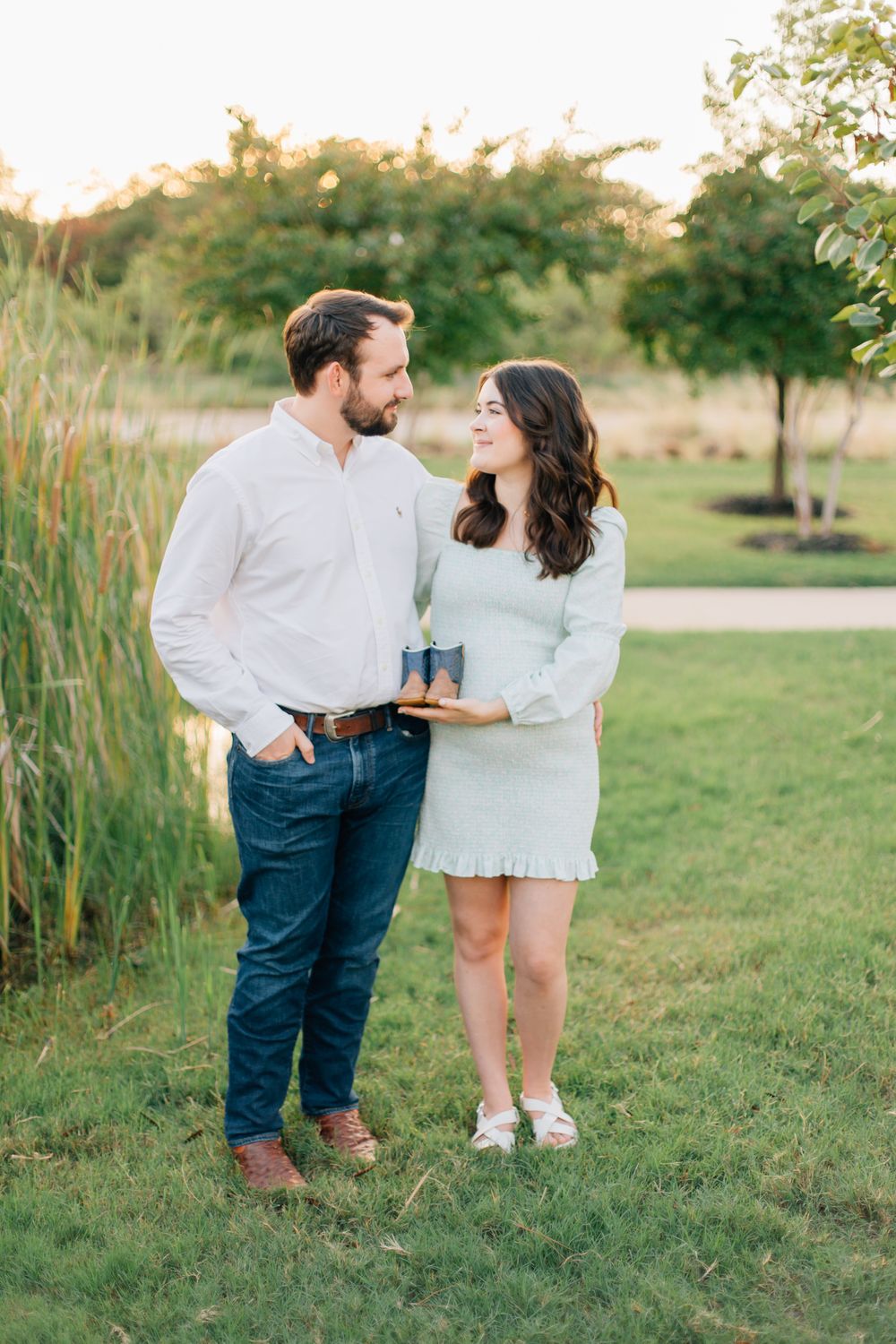 Happy couple shares an intimate moment outdoors on a grassy lawn at sunset with tall reeds in background.