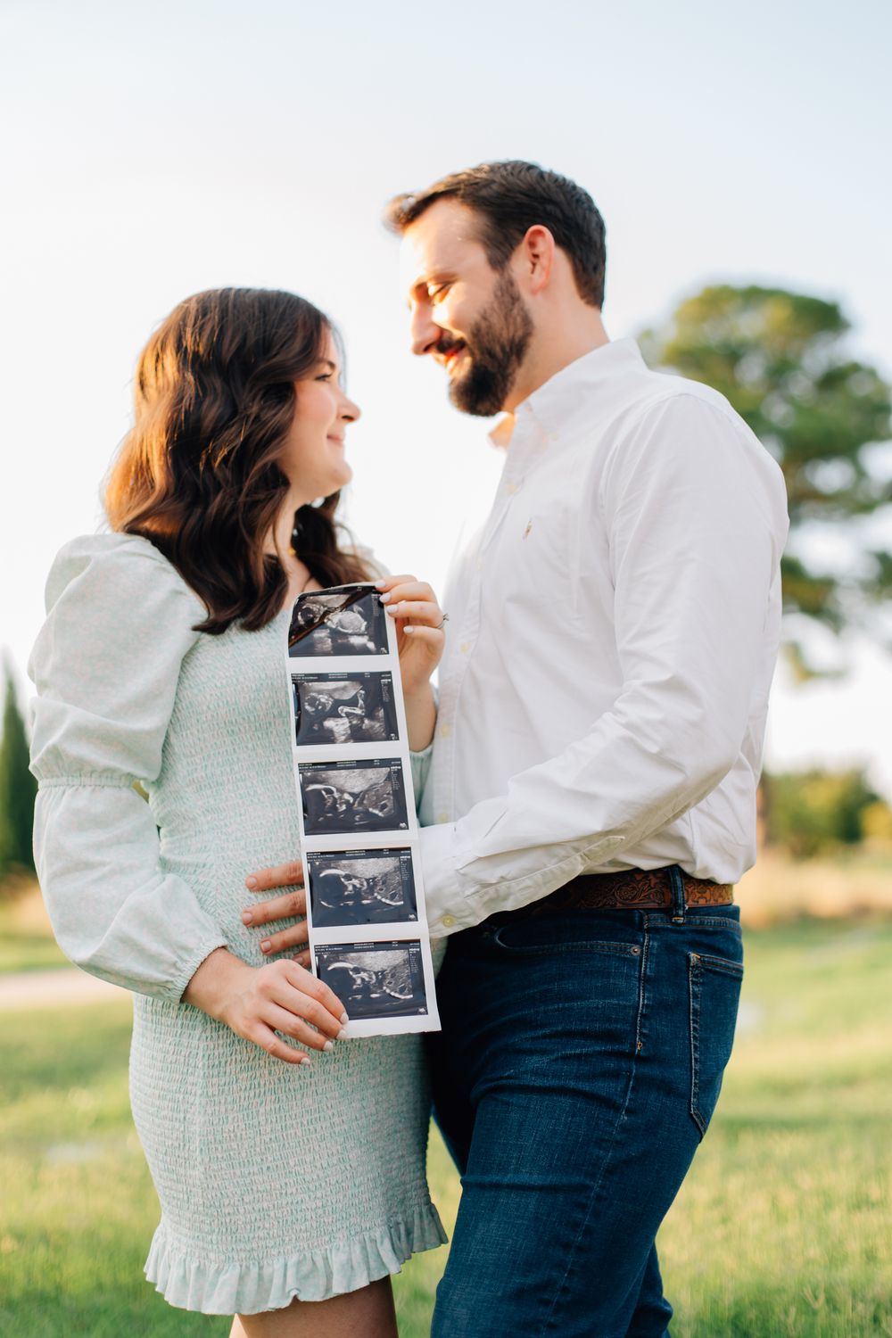 A joyful couple shares a tender moment outdoors while holding ultrasound photos in a pregnancy announcement photoshoot.
