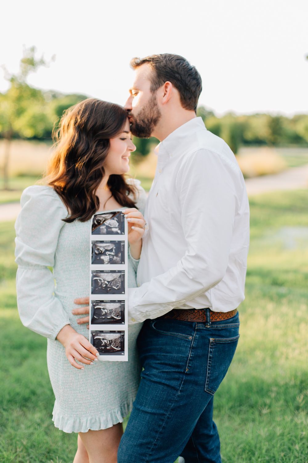 Couple in a sunlit field holding ultrasound photos celebrating pregnancy announcement during golden hour.