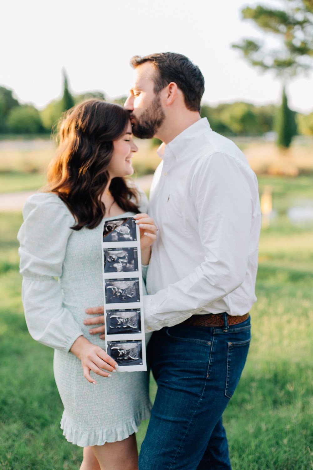 A couple stands close together outdoors holding ultrasound photos in a romantic pregnancy announcement photoshoot.