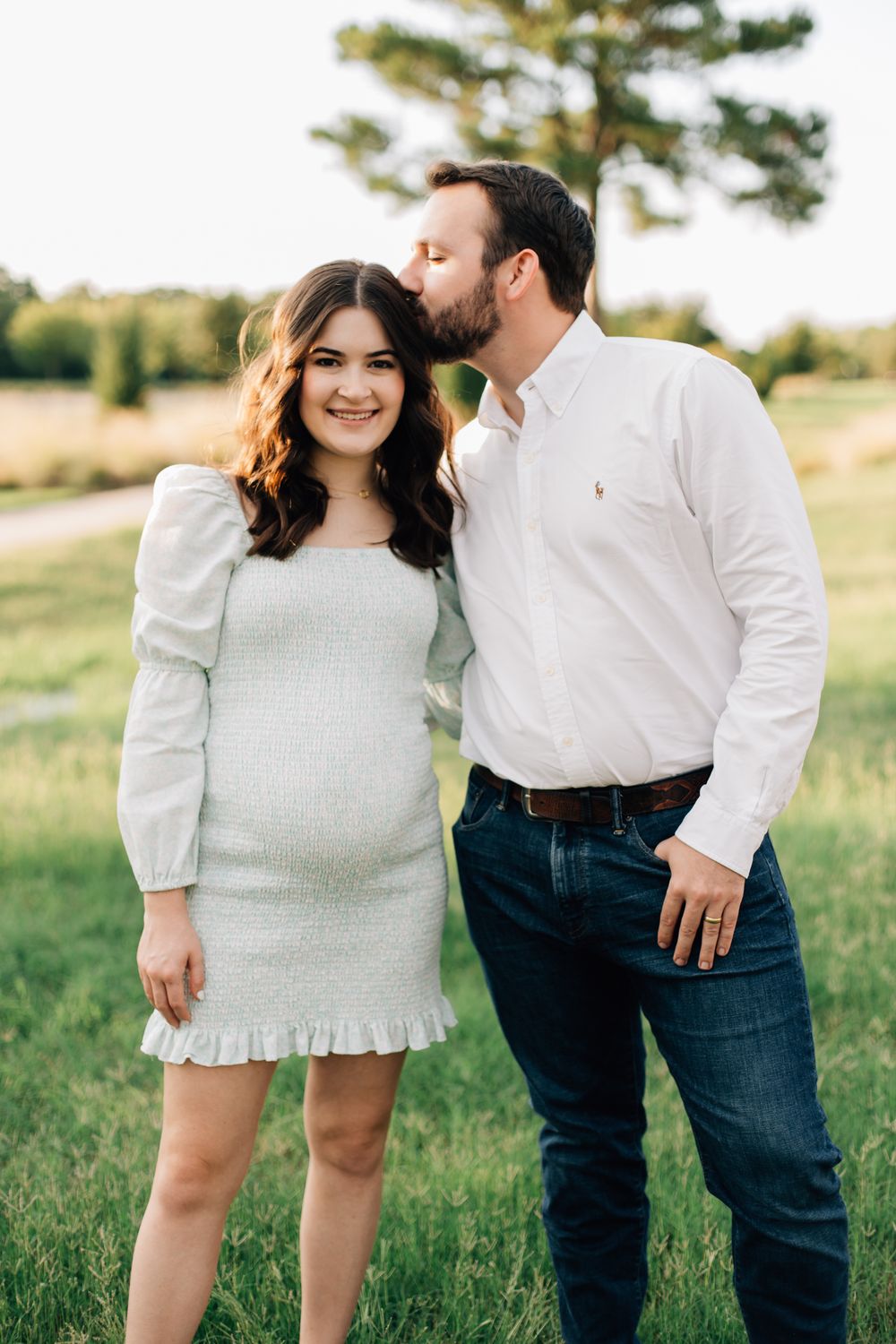A couple shares an intimate outdoor moment in a grassy field with a pine tree in the background.