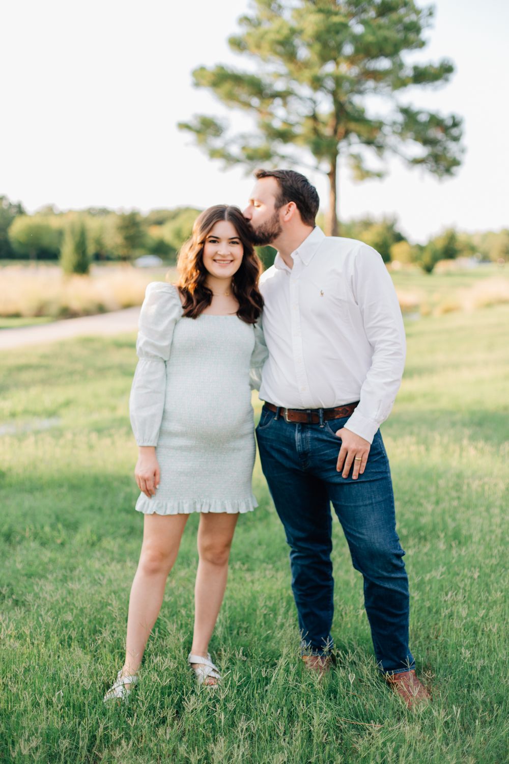 A couple stands together in a grassy field with pine trees in the background during a sunlit outdoor photoshoot.