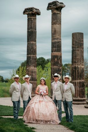 A quinceañera photoshoot featuring a pink ballgown and groomsmen in cowboy attire against ancient stone columns.