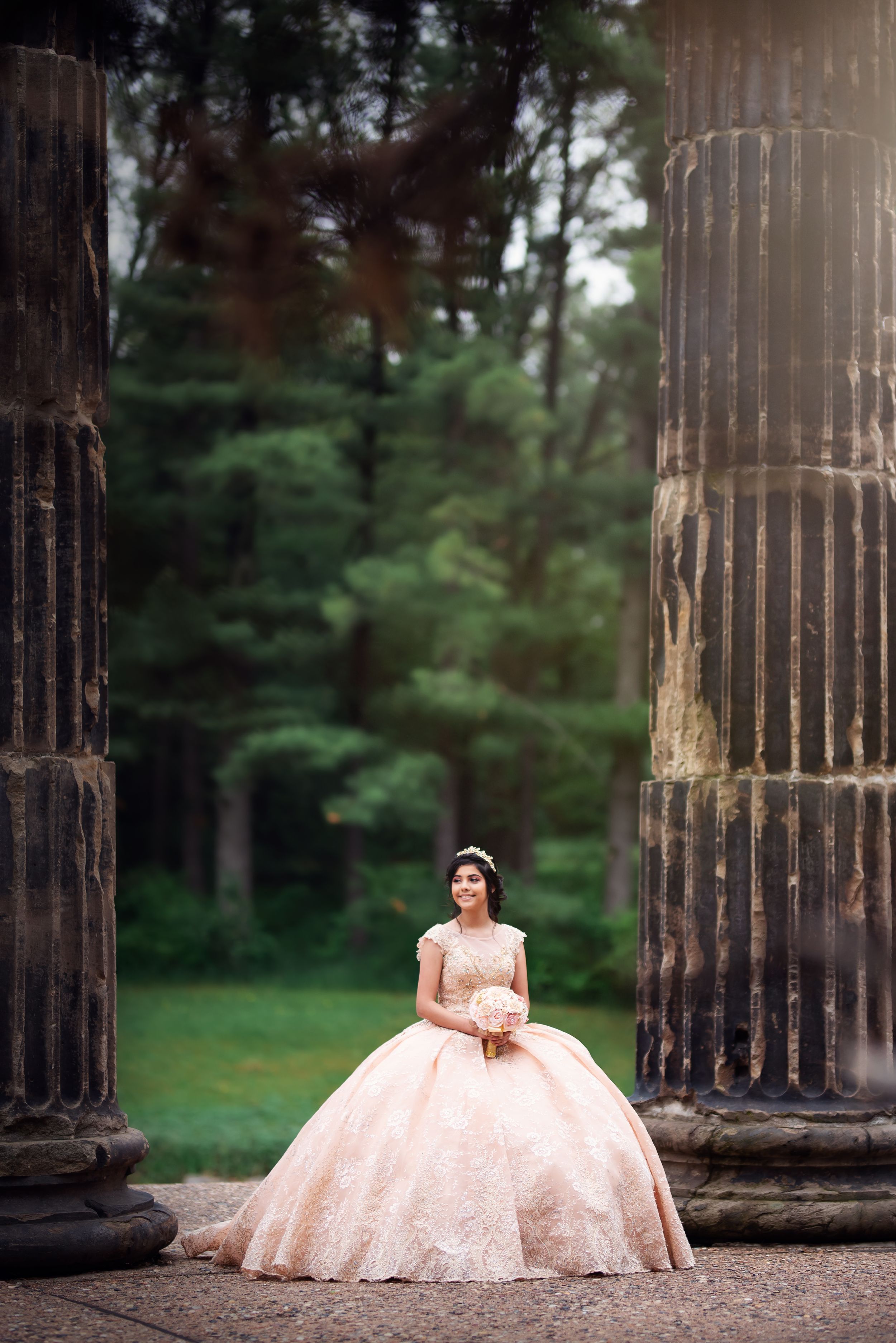 Elegant peachy-pink formal dress captured among classical stone pillars in a park setting.