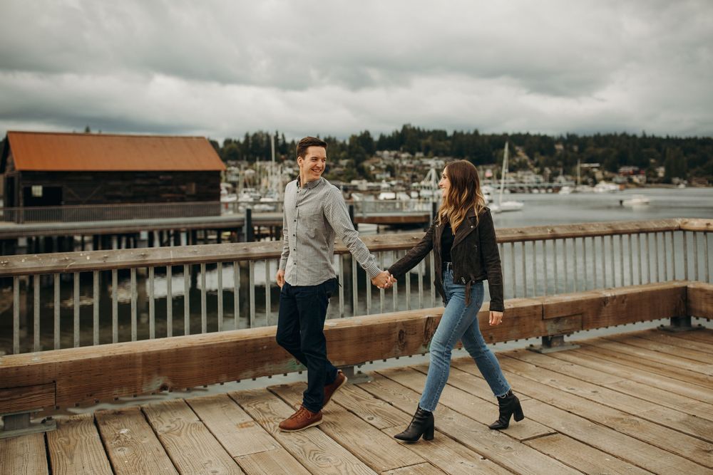 Two people walk and hold hands on a wooden dock overlooking a marina with boats and buildings in the background.