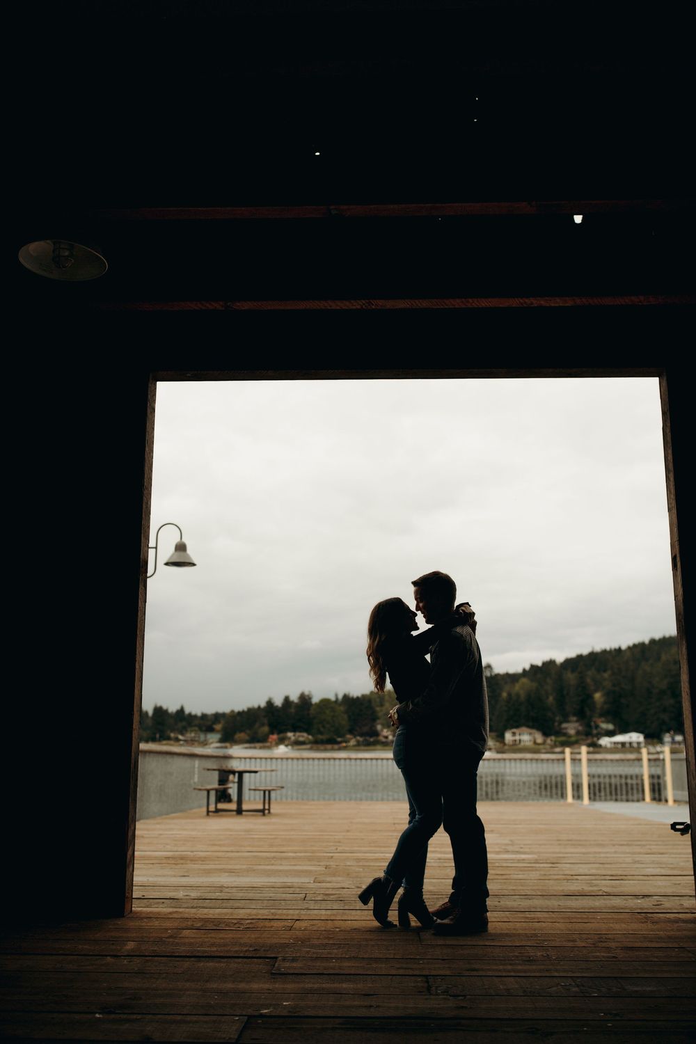Silhouetted couple embracing on wooden pier overlooking water and mountains at dusk framed by dark doorway.