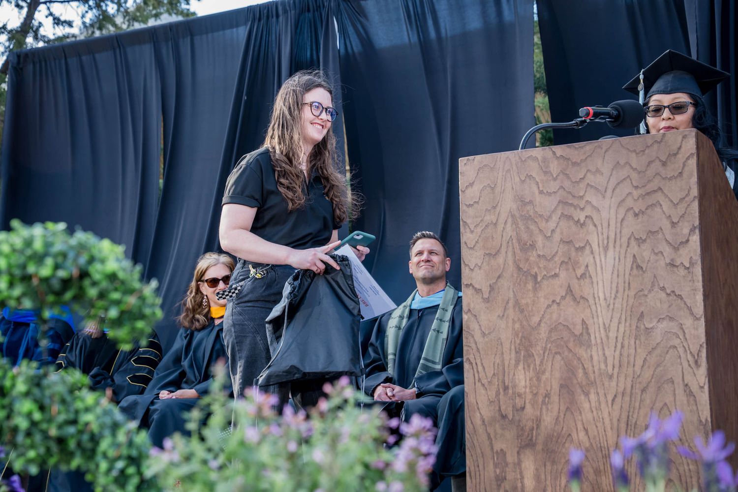 A graduation commencement ceremony with a presenter at a podium on stage, surrounded by purple flowers outdoors.