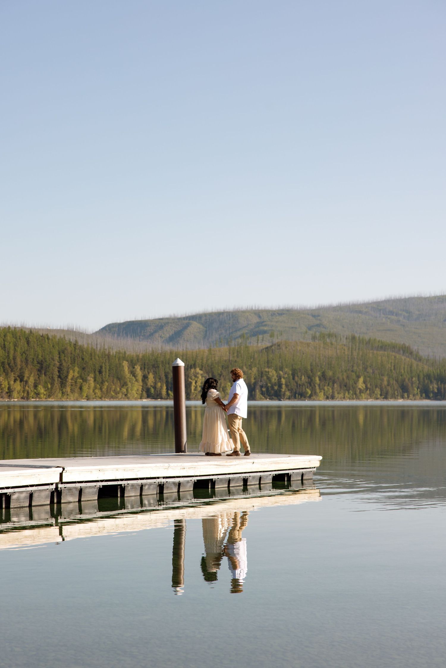 Couple stands together on wooden dock overlooking serene mountain lake with forest and hills in background.