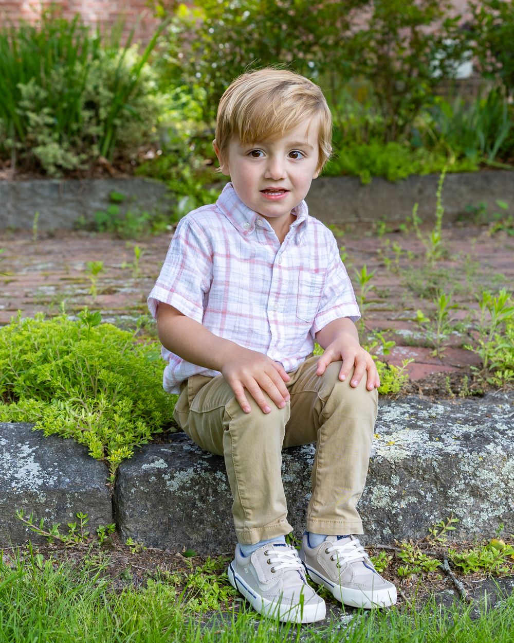 Young child in pink plaid shirt sitting on stone wall in garden setting during a family photo session in Sudbury, MA outside Boston.