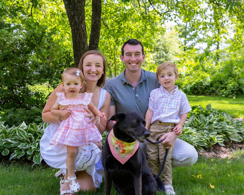 Family of four with black dog posing for portrait in lush garden setting during a family photo session in Sudbury, MA outside Boston.