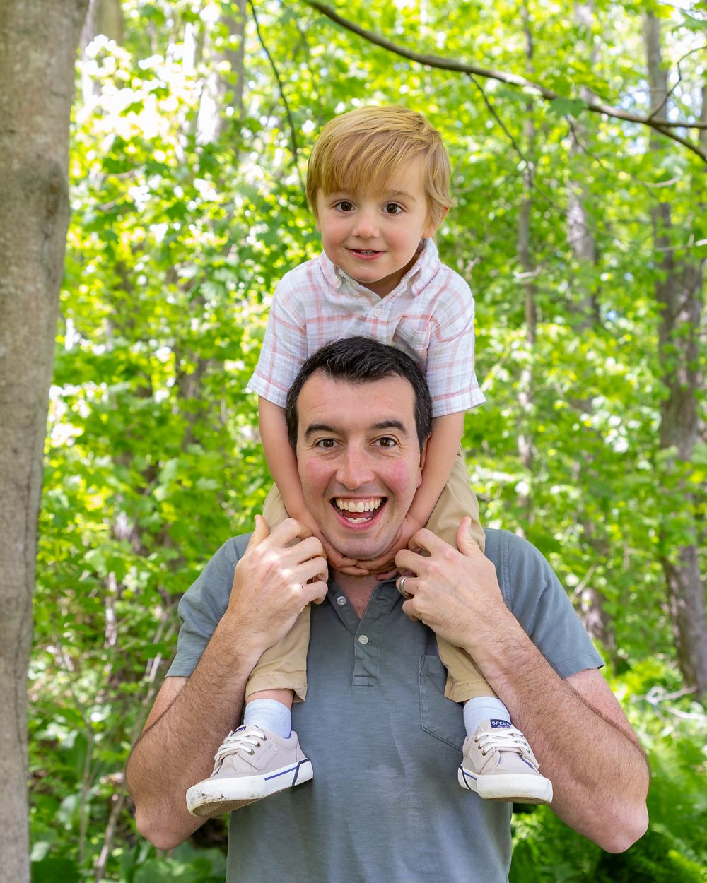Playful portrait of child riding on shoulders in wooded outdoor setting during a family photo session in Sudbury, MA outside Boston.