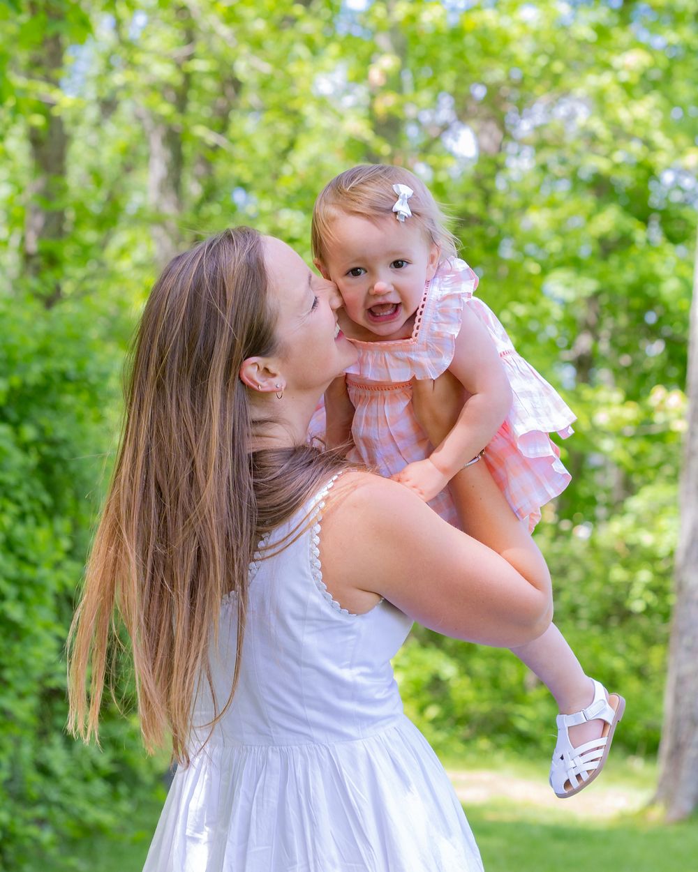 A sweet outdoor portrait of mother and baby in matching white dresses amid lush green foliage on a sunny day in Sudbury, MA