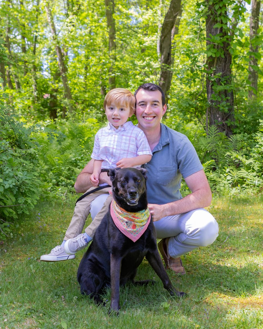 A family poses outdoors in a lush green forest setting with their black Lab mix dog wearing a bandana at a portrait session in Sudbury, MA