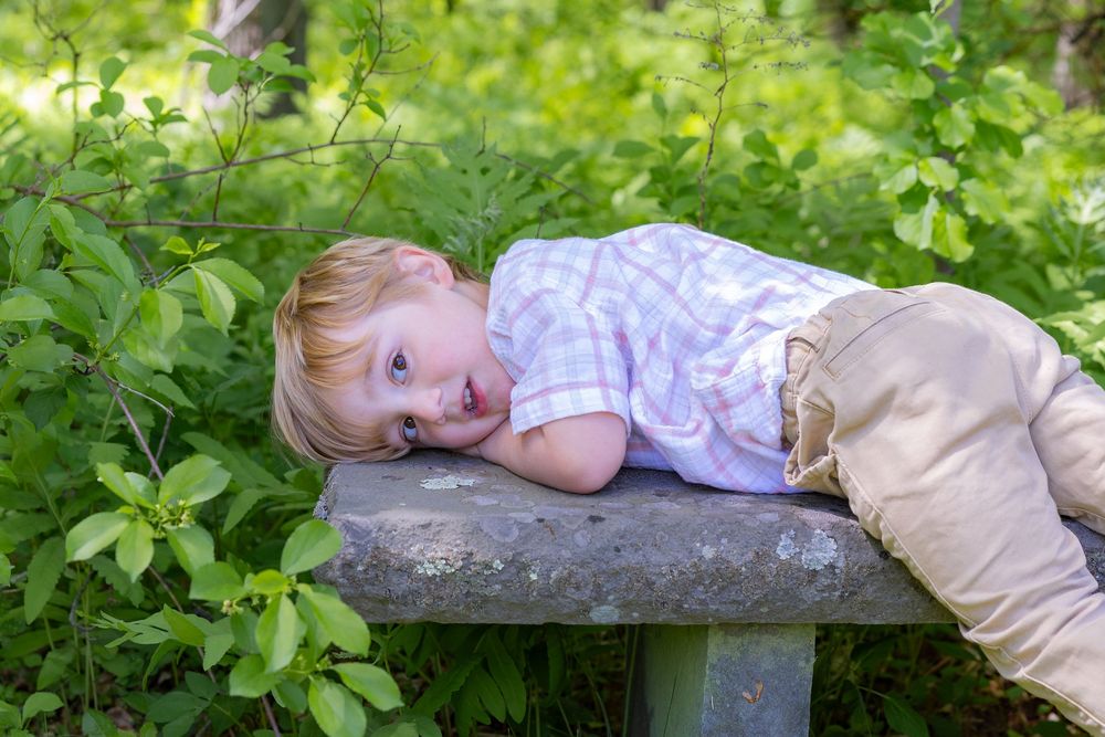 A toddler boy on a stone bench surrounded by lush green foliage in a garden setting during a family photo session in Metrowest of Boston.