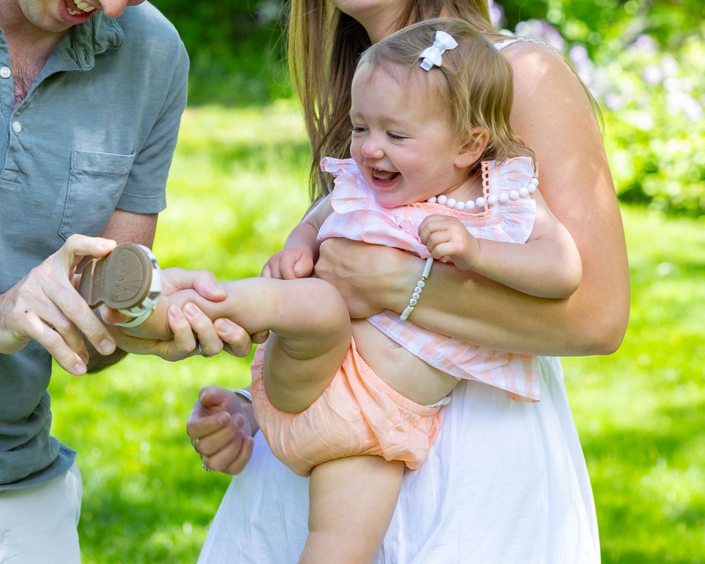 Laughing toddler in pink dress being tickled while sitting on lap in sunny garden setting during summer during a family photo session