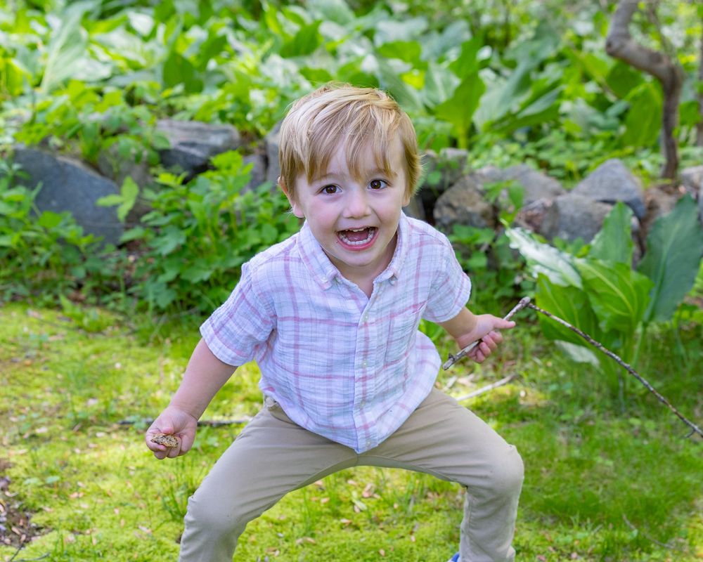 Energetic young child in plaid shirt striking playful pose in garden setting during a family photo session in Sudbury, MA outside Boston.