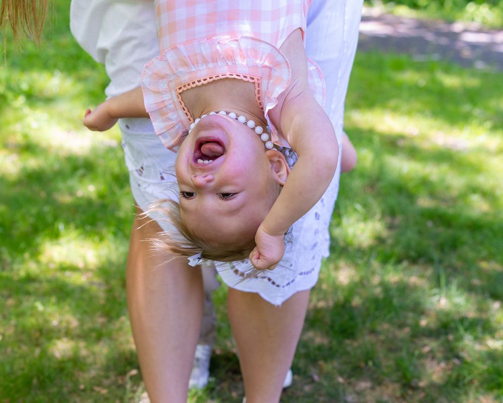 Playful upside-down baby in pink dress laughing during outdoor portraits during a family photo session in Sudbury, MA outside Boston.