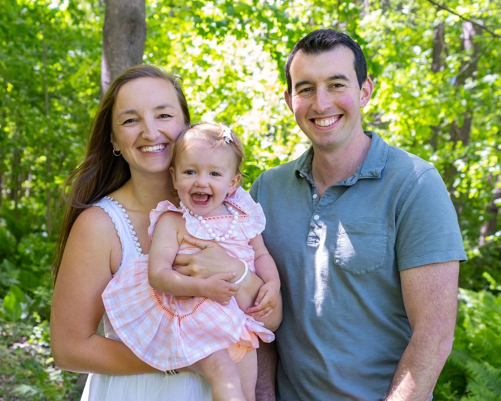 Cheerful family portrait outdoors with baby in pink dress surrounded by green foliage during a family photo session in Sudbury, MA