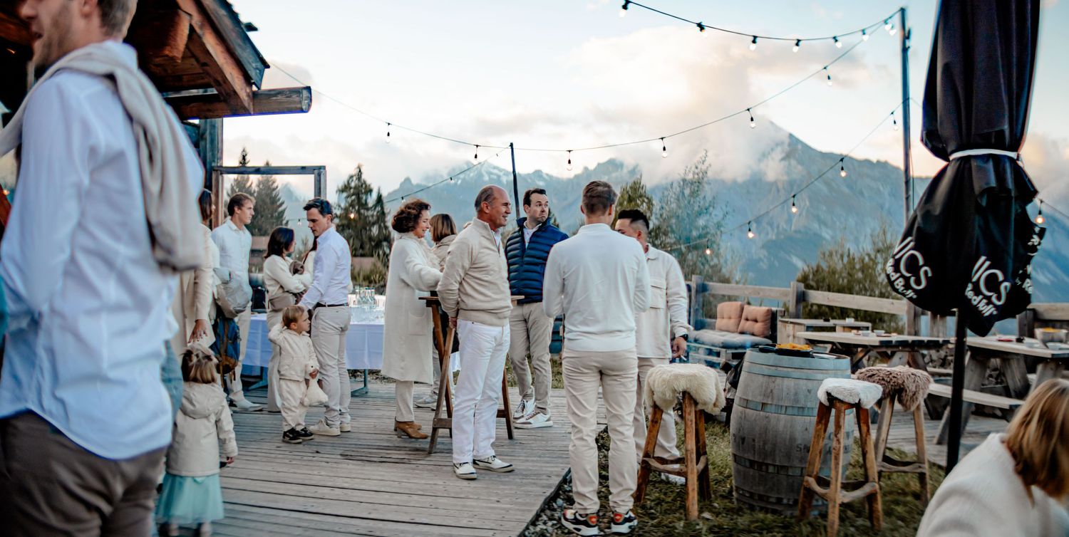 People gather at an outdoor mountain venue deck with string lights and white attire for an elegant evening celebration.