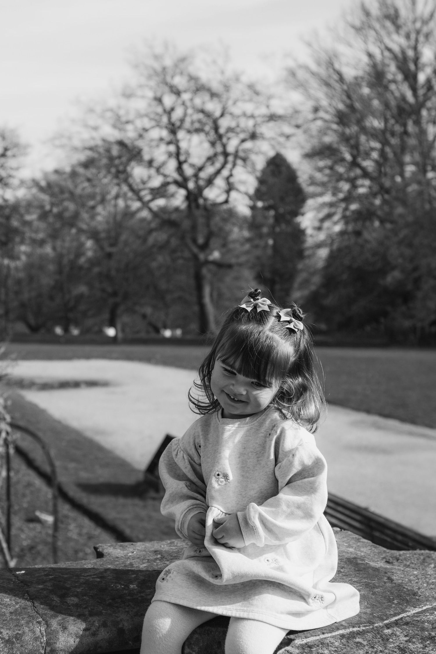 Black and white photograph of a young child in a light dress sitting on a stone wall in a park with trees in background in Nottingham