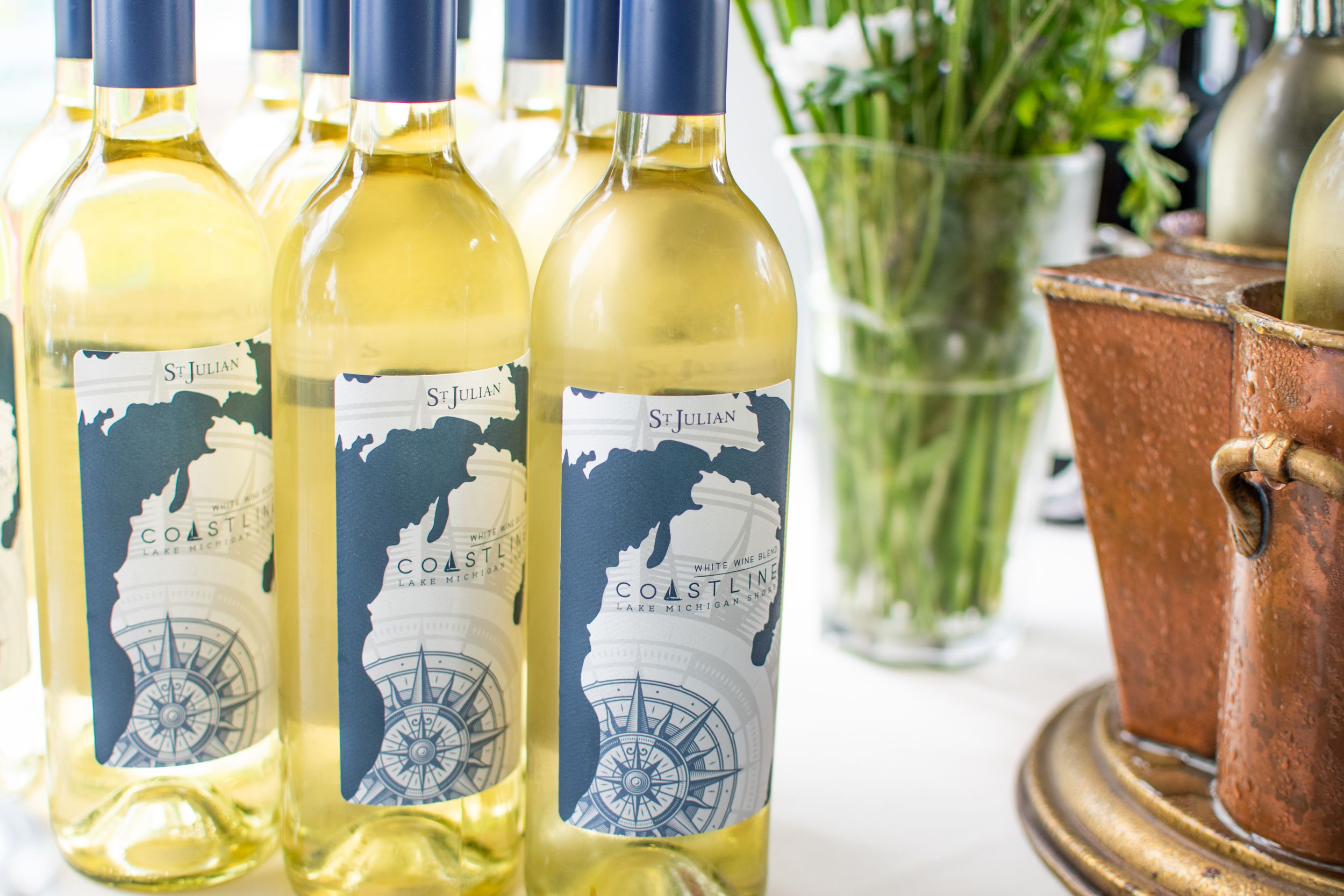 Three bottles of white wine with custom nautical-themed labels displayed near potted plants.
