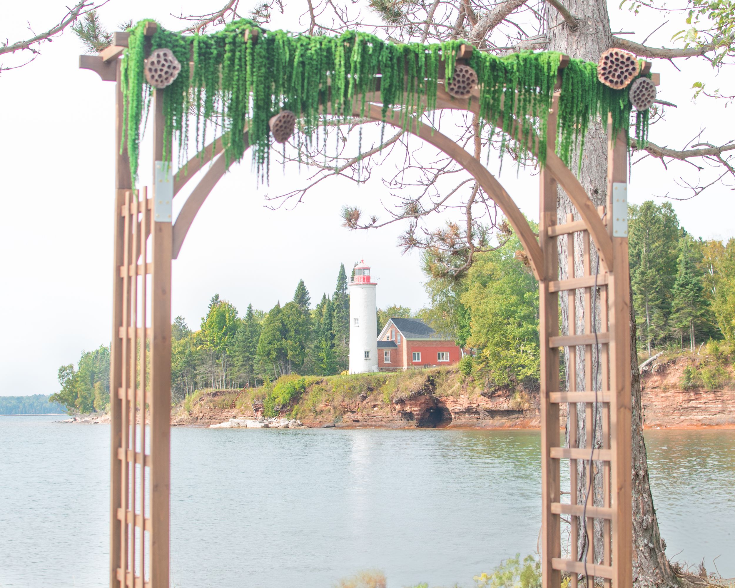 A wooden wedding arch draped with greenery frames a lighthouse on a rocky shoreline.