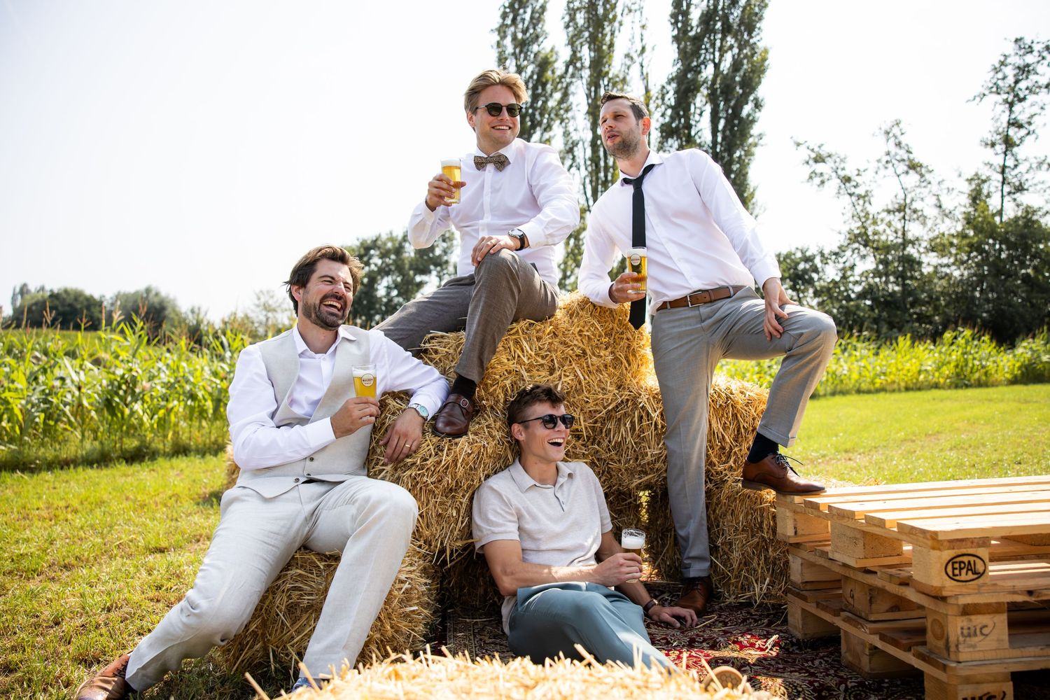 A group in matching outfits poses casually on hay bales in a sunny outdoor setting with trees in background.