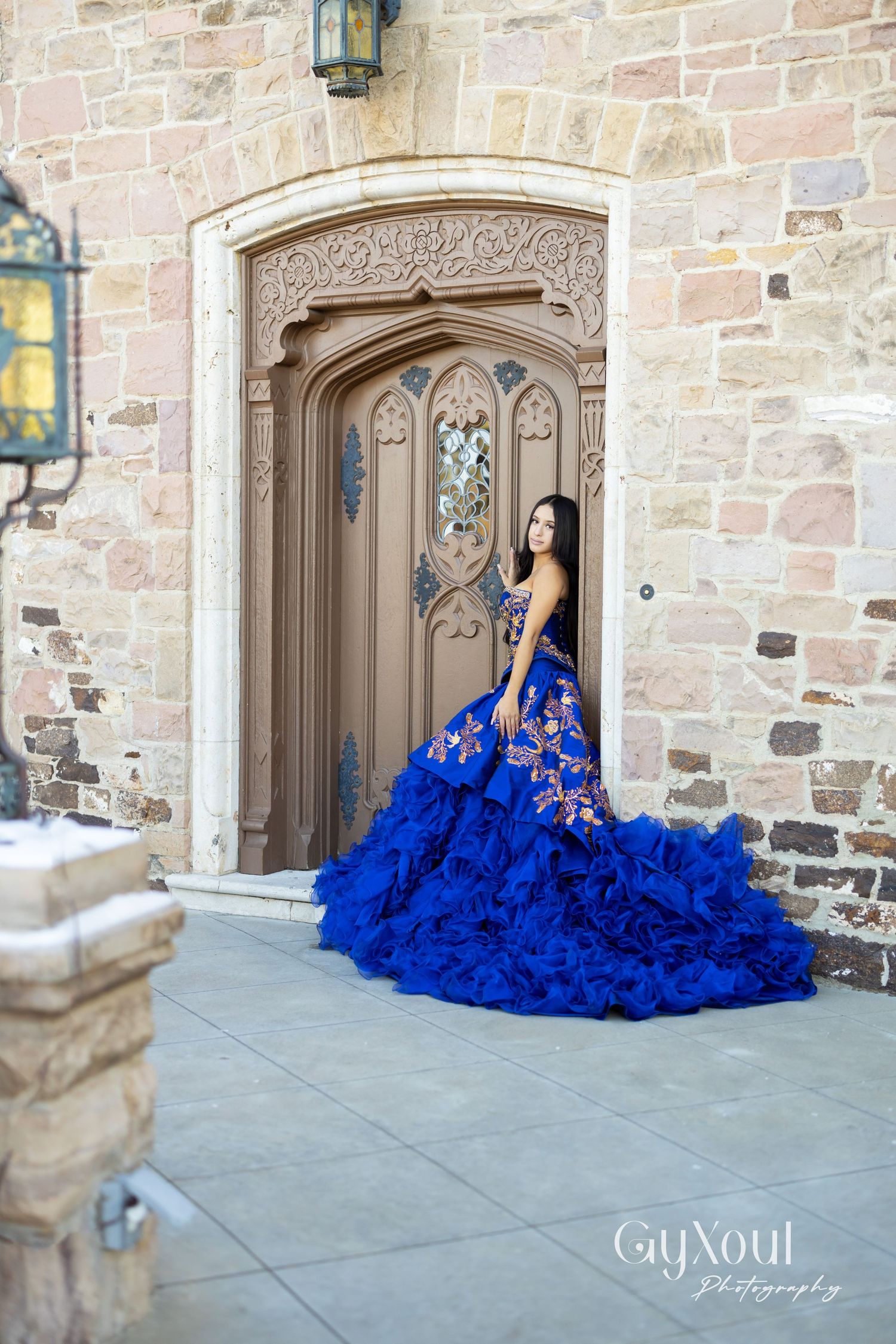 Elegant royal blue quinceañera dress with ruffled train photographed in front of ornate stone archway and wooden door.