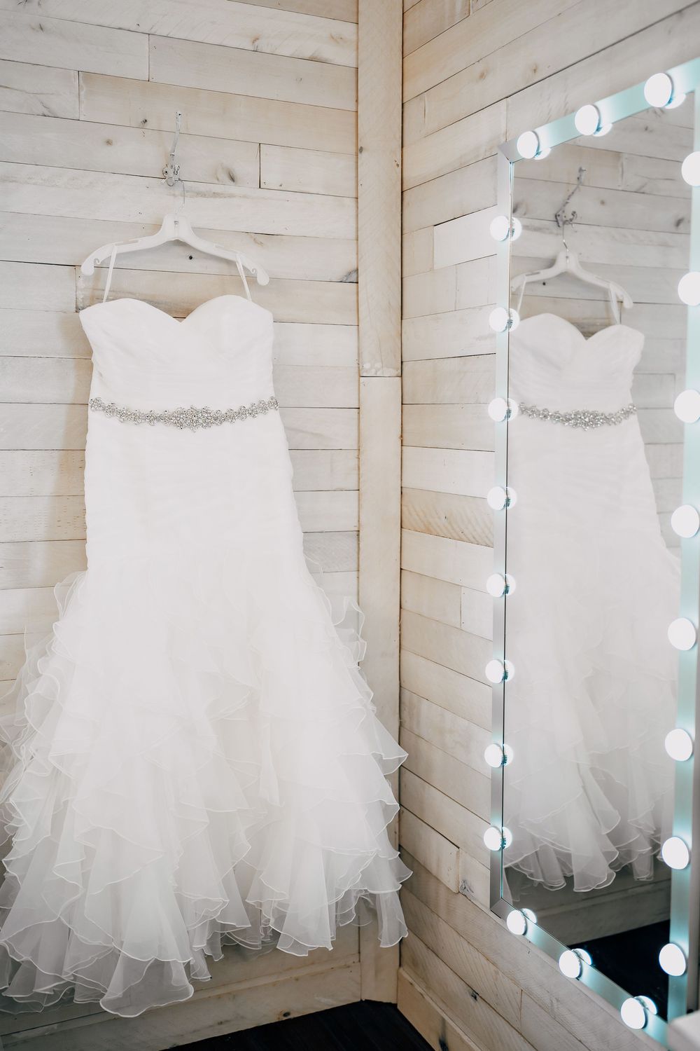 A beautiful white wedding dress with tulle skirt and beaded belt hangs next to a mirror with vanity lights on wooden walls.