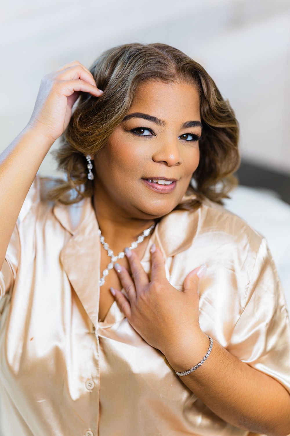 Bride in silk champagne robe wearing pearl jewelry touches her softly curled hair and smiles.