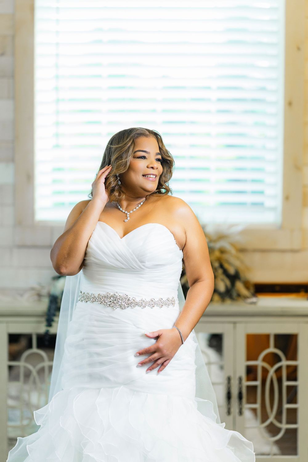 Beautiful bride in a white strapless gown with crystal belt poses by a bright window.