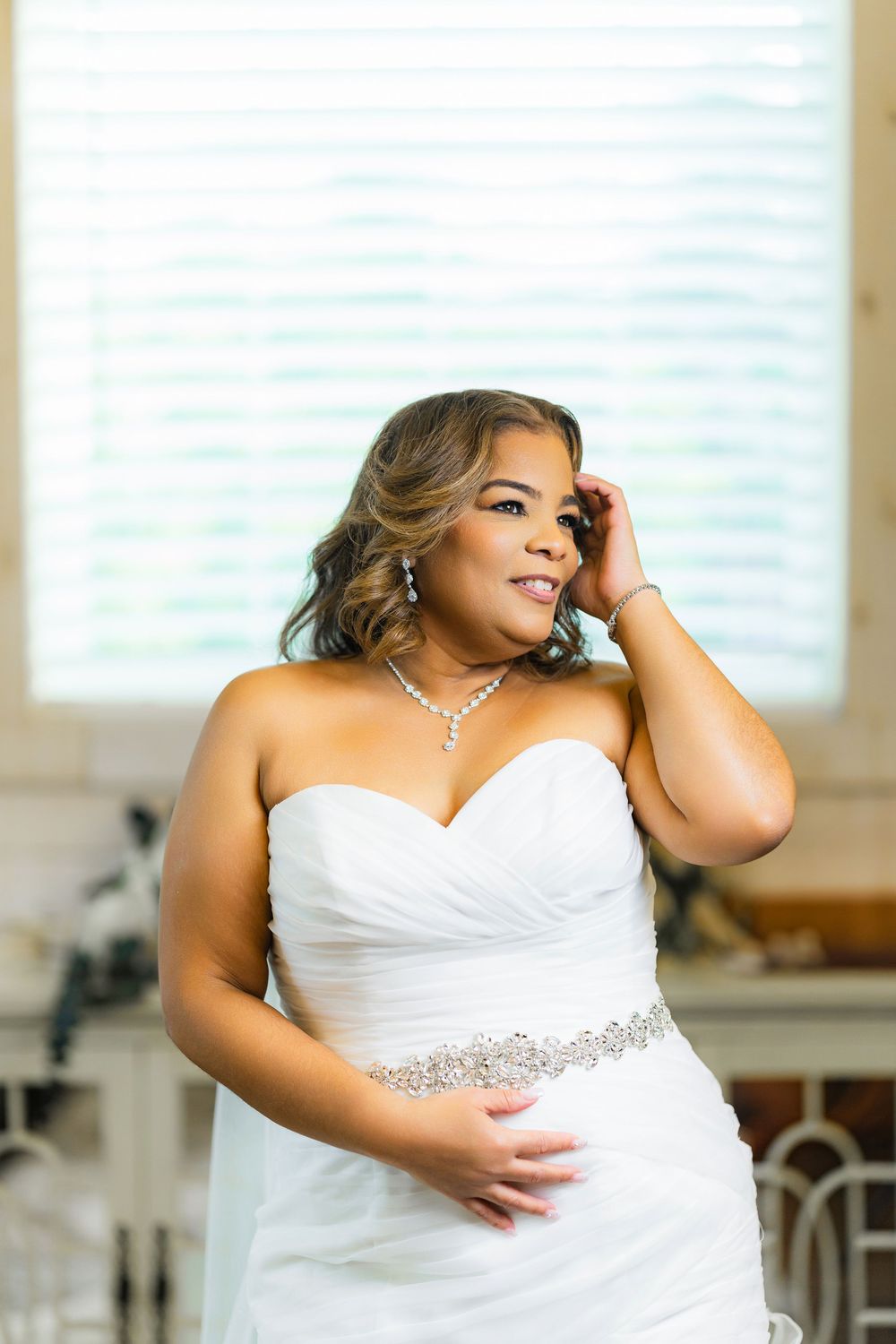 Person wearing white strapless wedding gown with crystal belt detail in sunlit room.