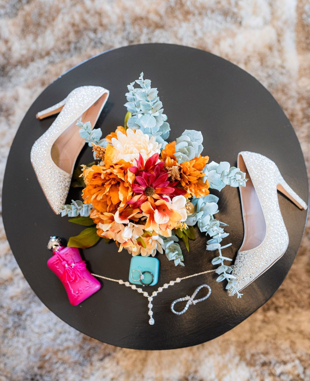 Wedding details with white sparkly heels and vibrant orange and blue floral bouquet arranged on a dark surface.