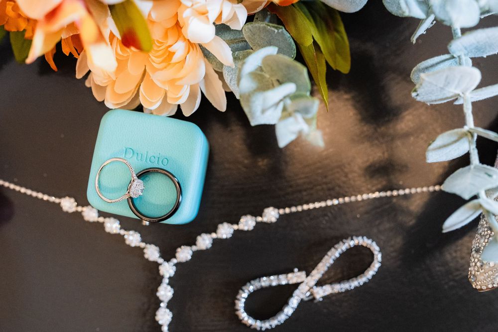Wedding details including sparkly jewelry, an infinity brooch, and teal ring box arranged with peach flowers and eucalyptus.