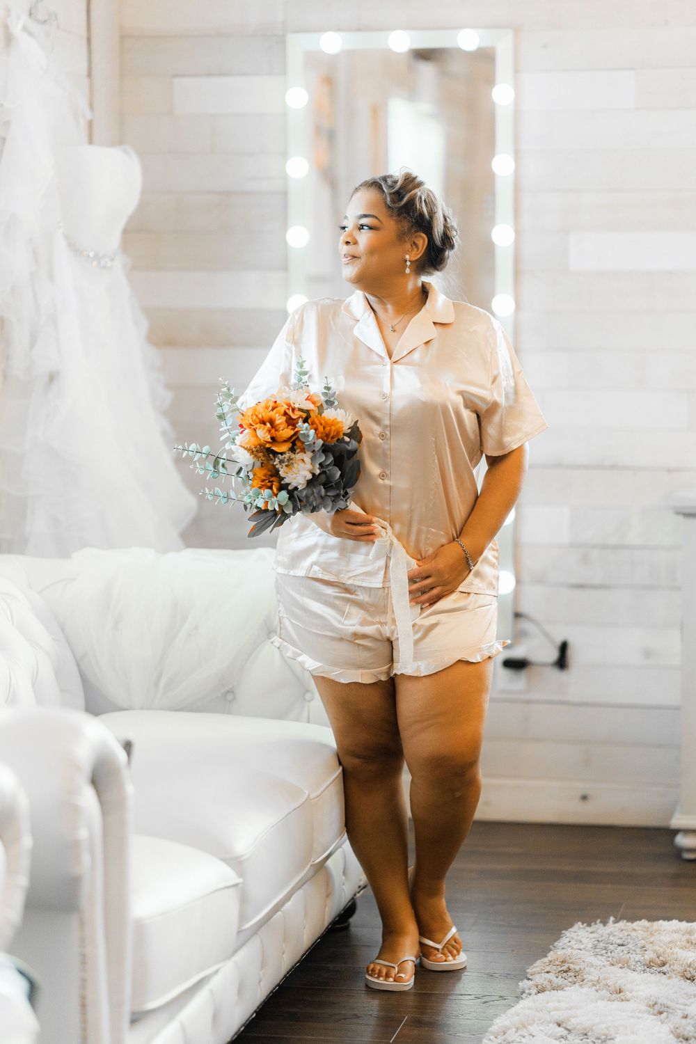 A person wearing silk pajamas holds an orange and eucalyptus bouquet while standing in a bright room with a mirror.