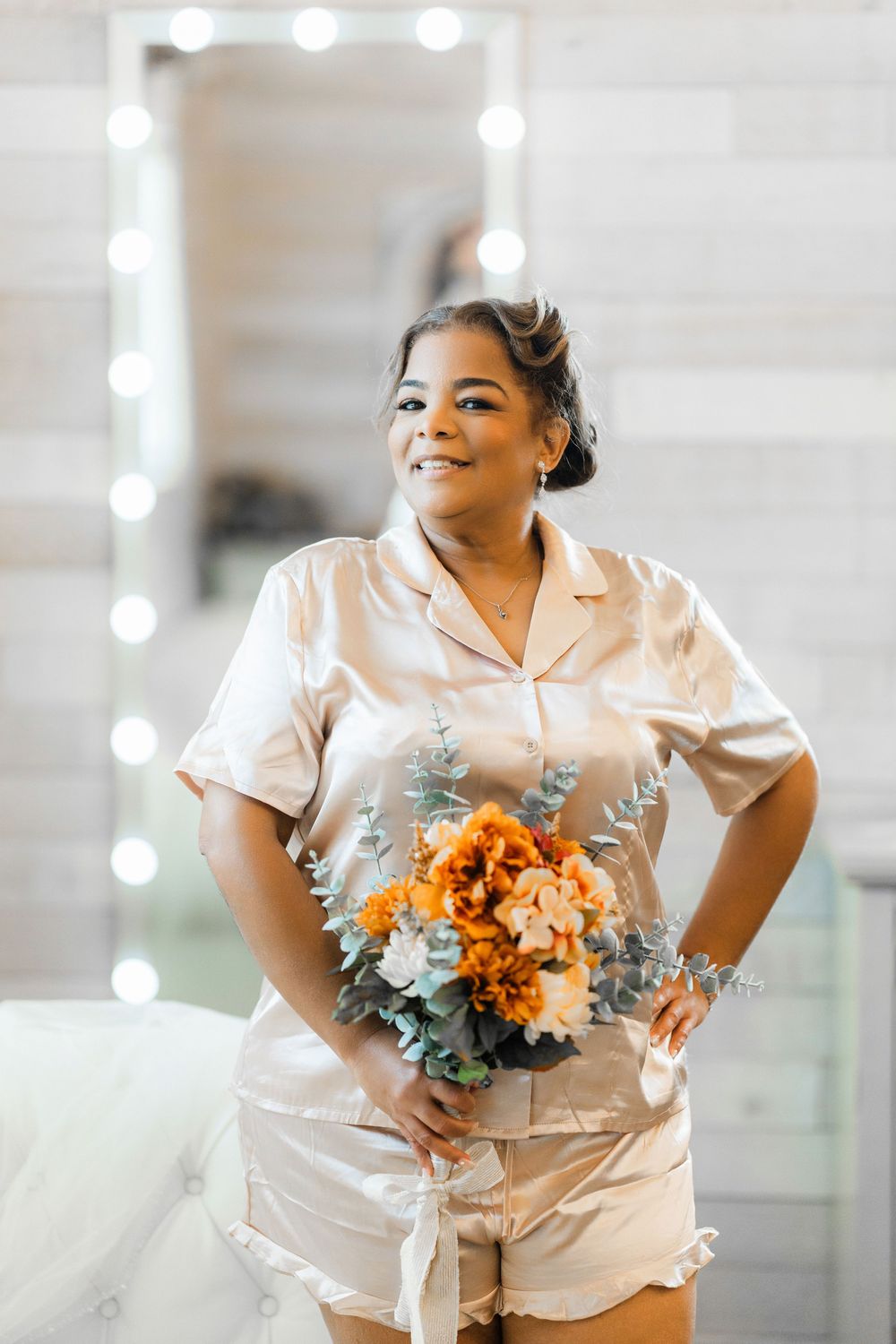 A person in silk sleepwear poses with an orange floral bouquet against a light-filled backdrop with a vanity mirror.