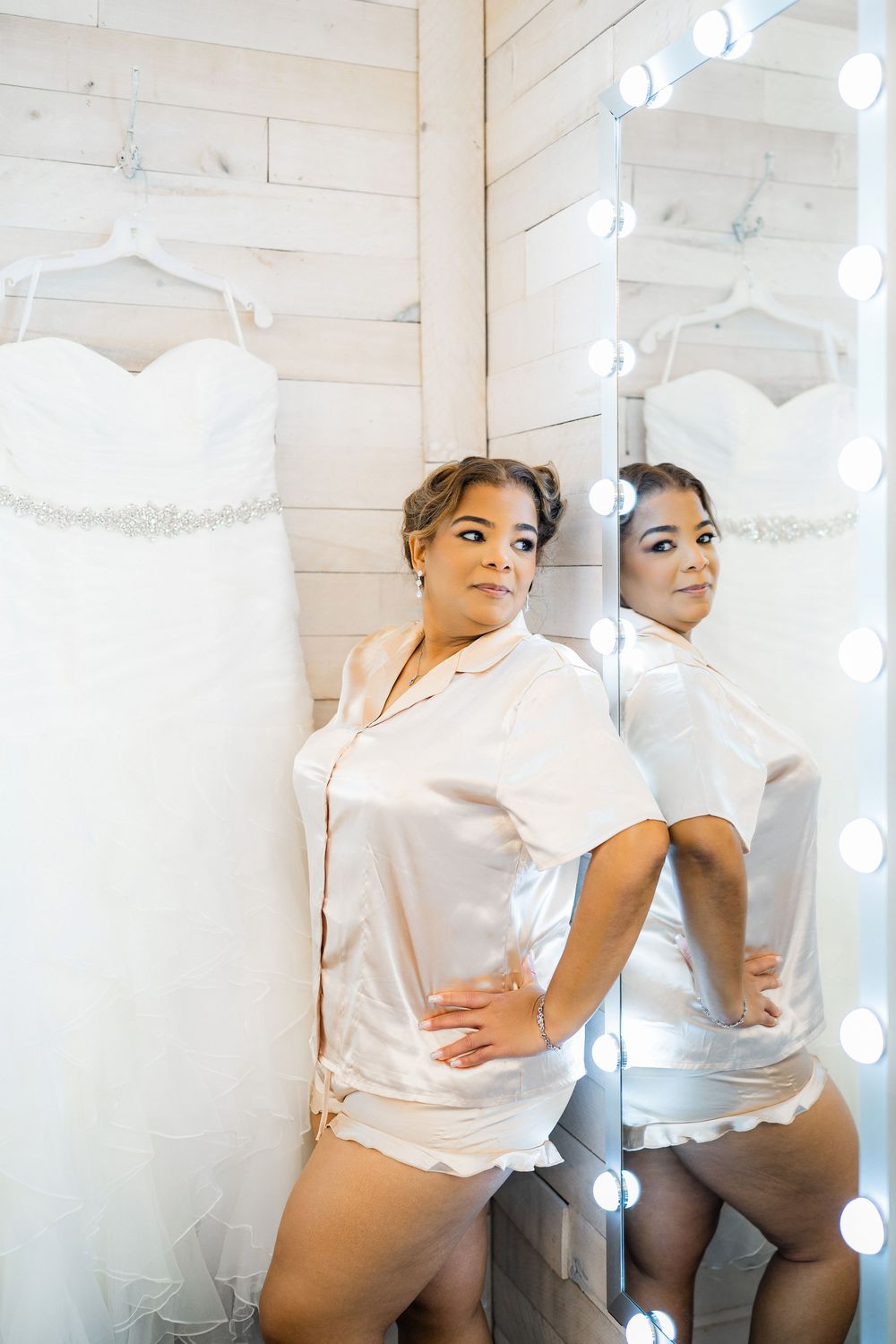 Wedding dress displayed on wall with person in white shirt looking in illuminated mirror with curled hair.