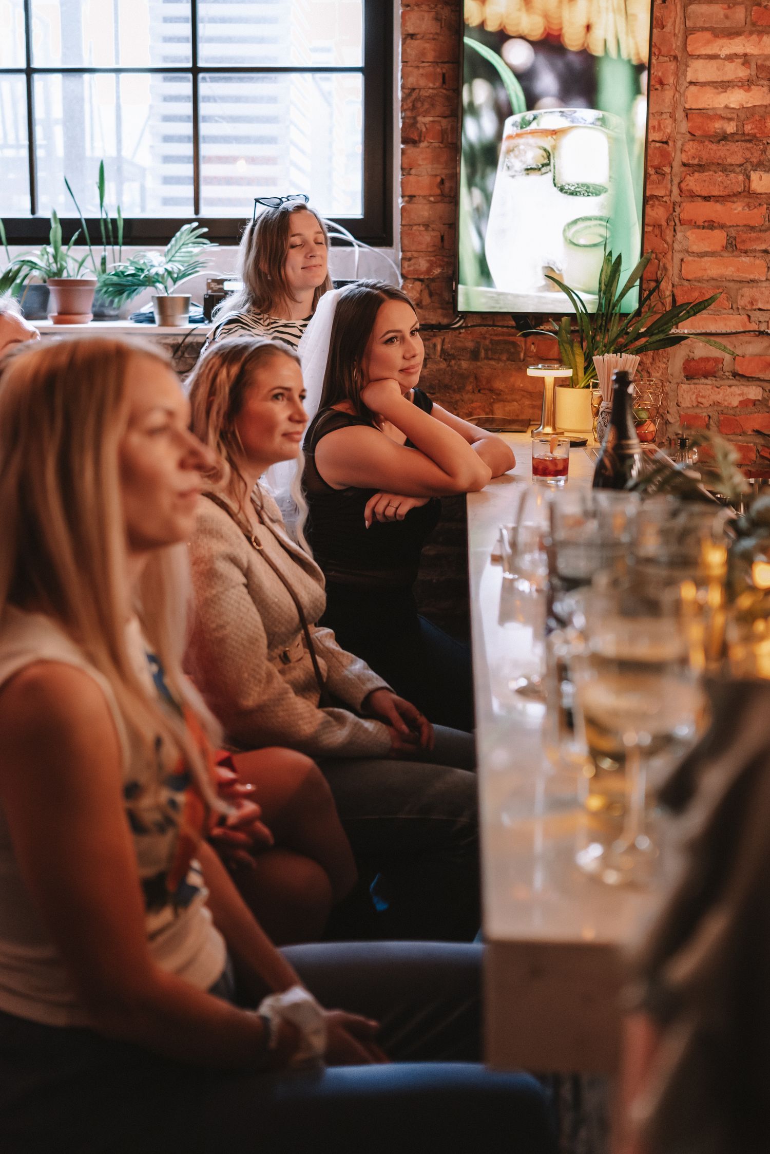 A group of customers sitting at a bar counter inside a rustic brick-walled establishment with mood lighting.