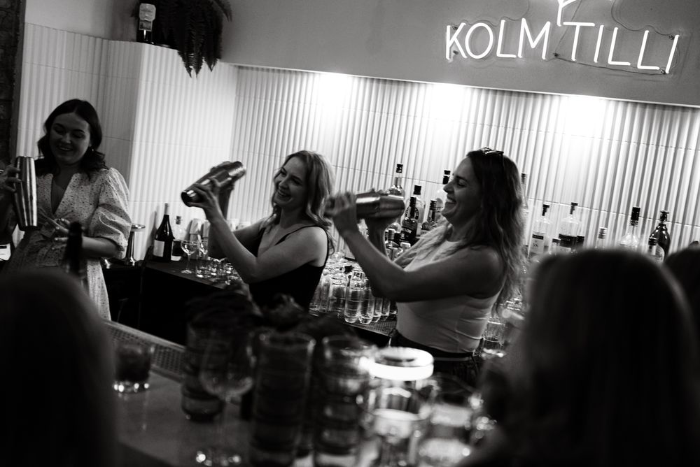 Party guests raise their glasses for a toast under illuminated 'KOLM TILLI' signage in a dimly lit setting.