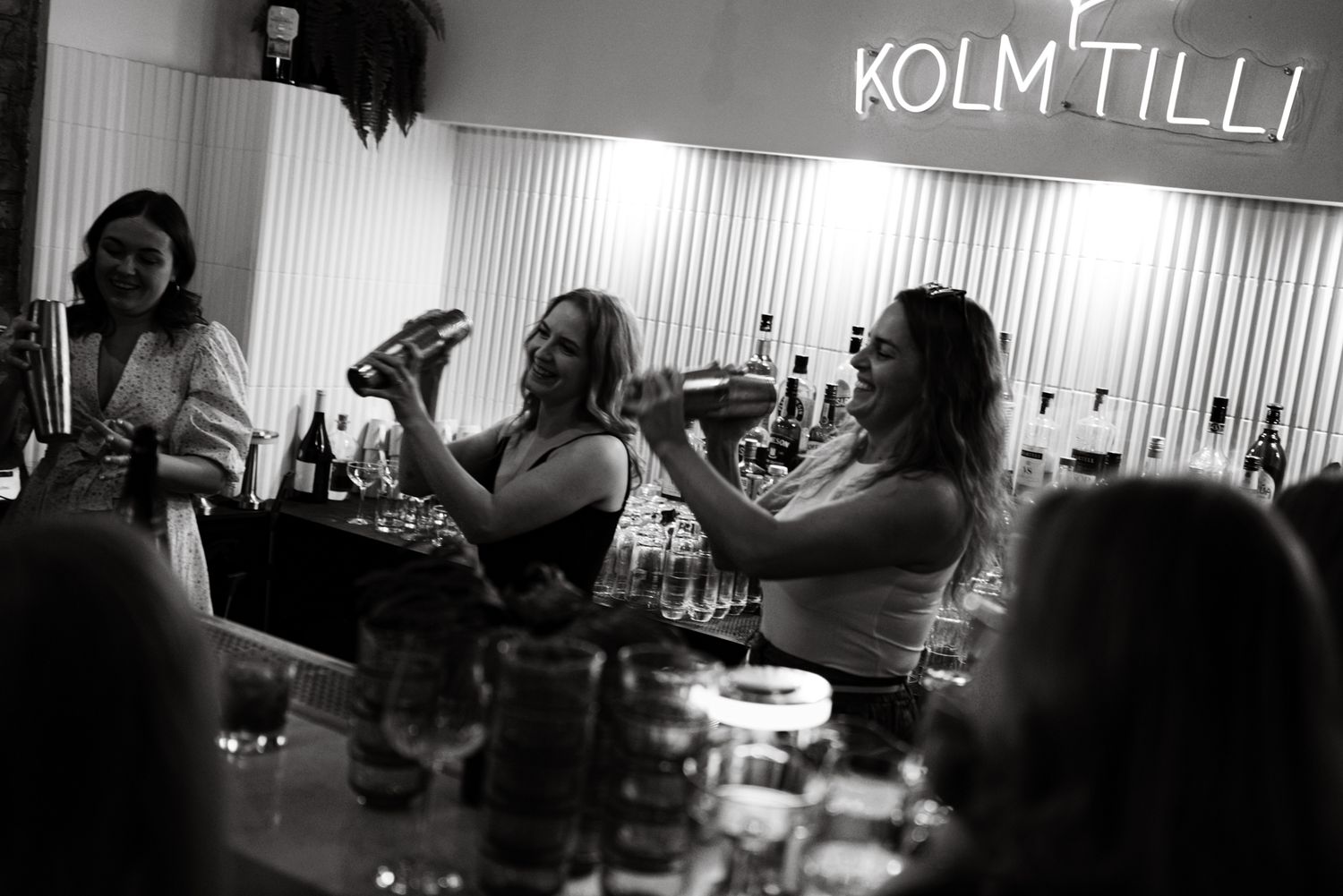 Party guests raise their glasses for a toast under illuminated 'KOLM TILLI' signage in a dimly lit setting.