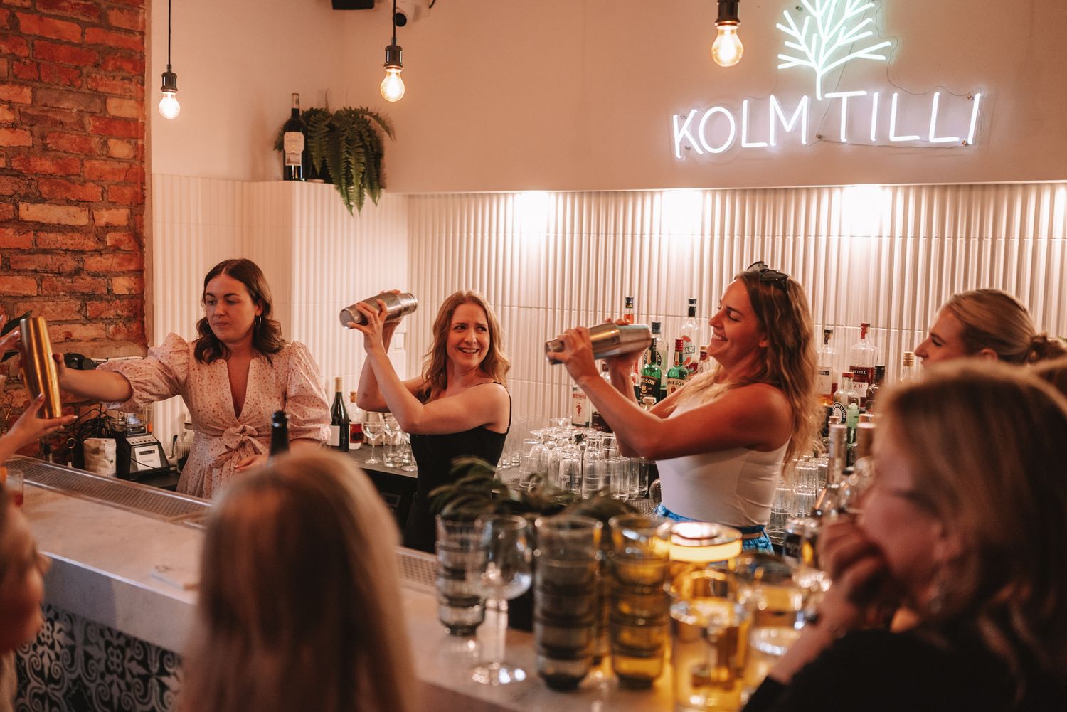 Group enjoying drinks and laughter at a trendy bar with neon Kolm Tilli sign and exposed brick walls.