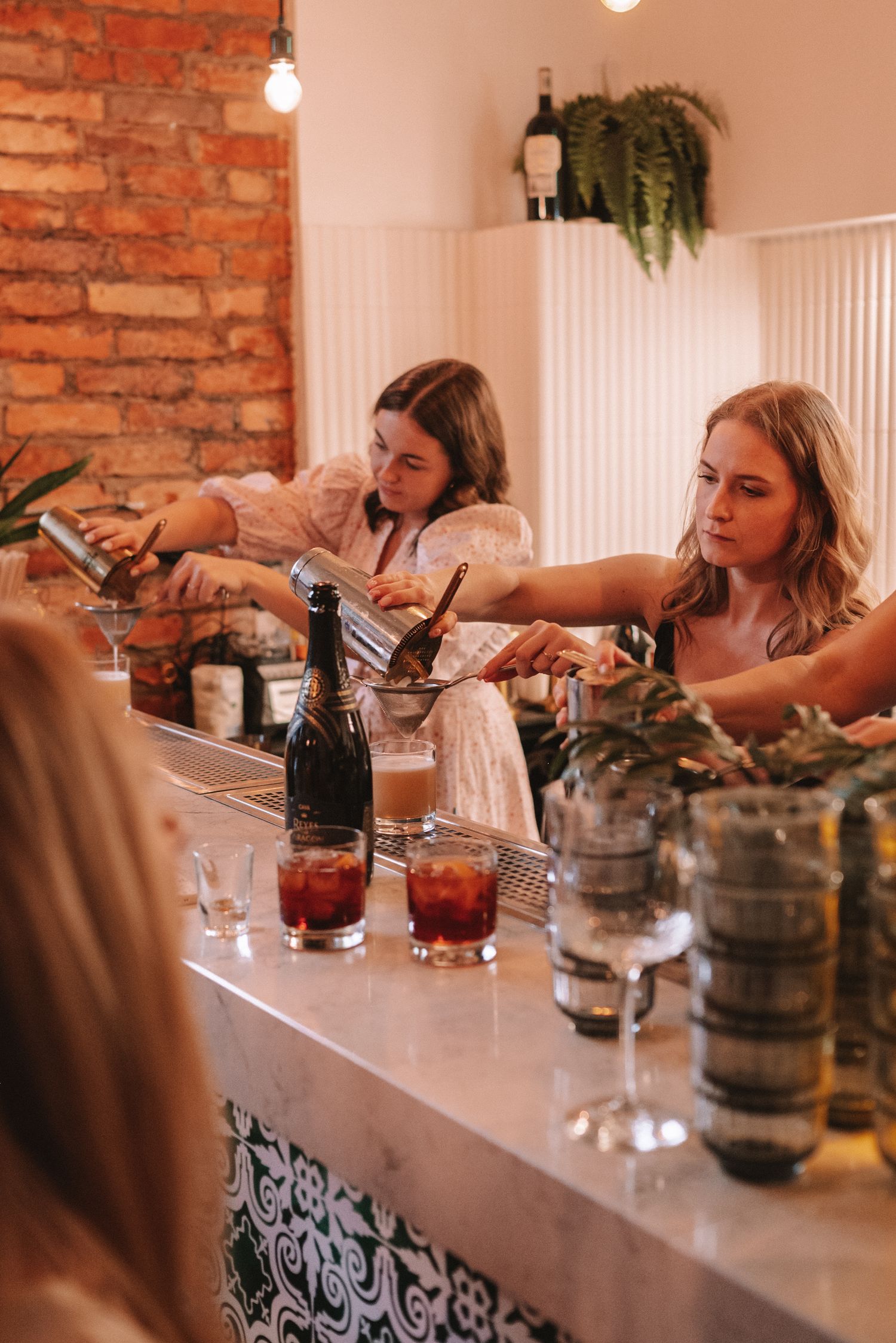 Bartenders prepare cocktails behind a bar counter with exposed brick walls and hanging plants in warm lighting.