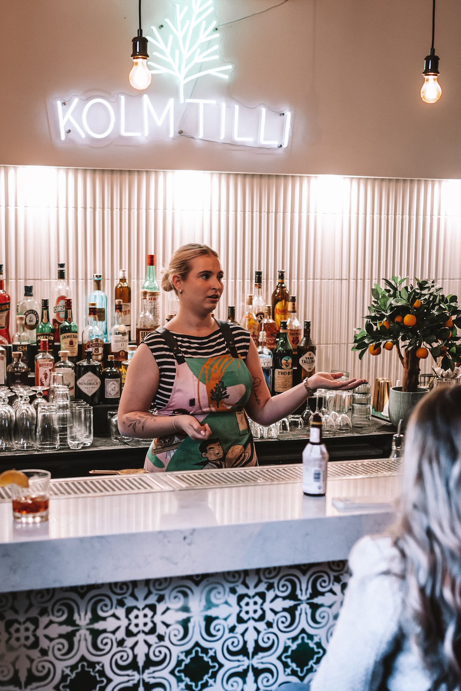 A bartender works behind a stylish bar counter underneath a glowing neon 'Kolmtill' sign with bottles displayed on shelves.