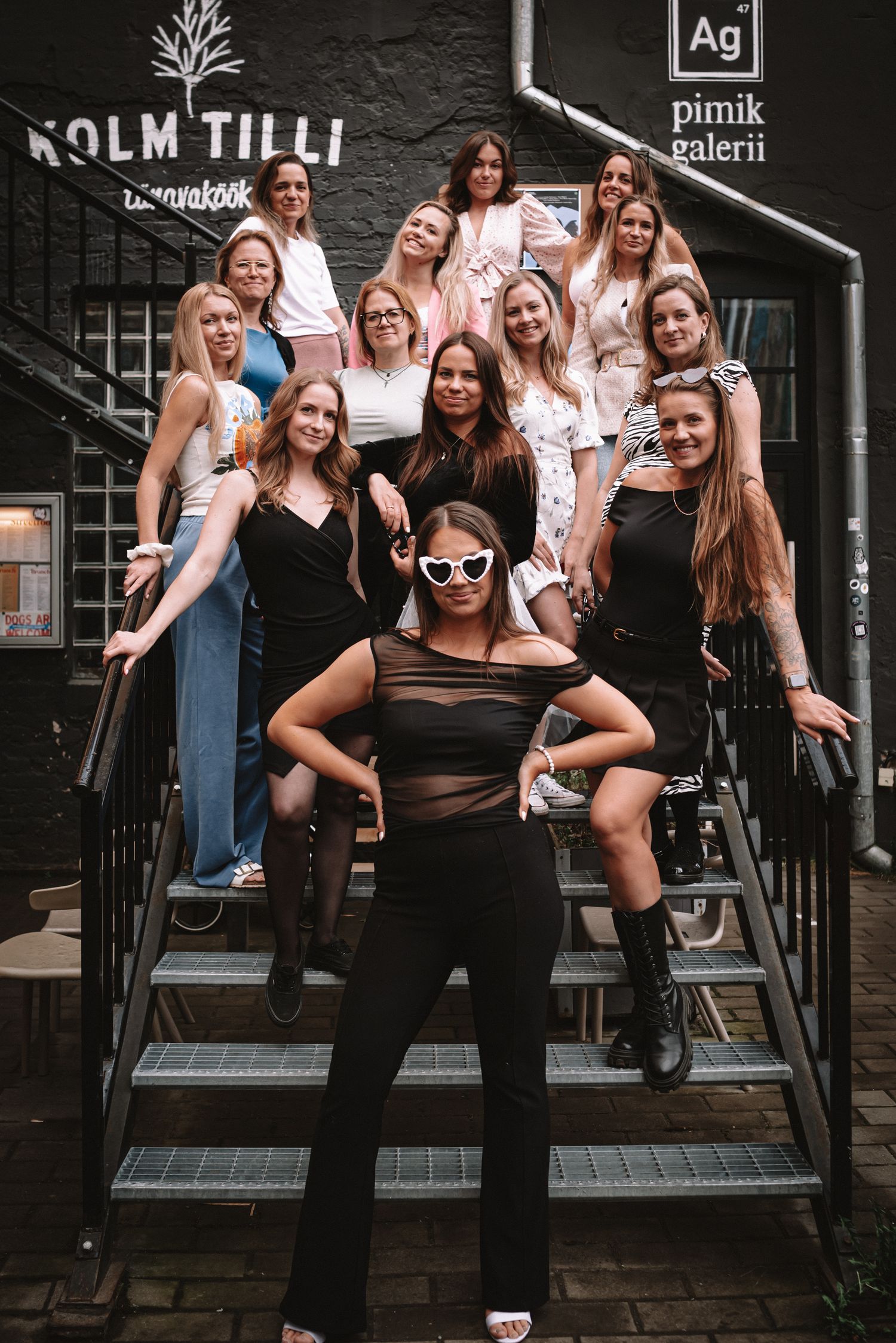 A group poses together on an outdoor staircase in front of a dark storefront, wearing a mix of black and light-colored outfits.