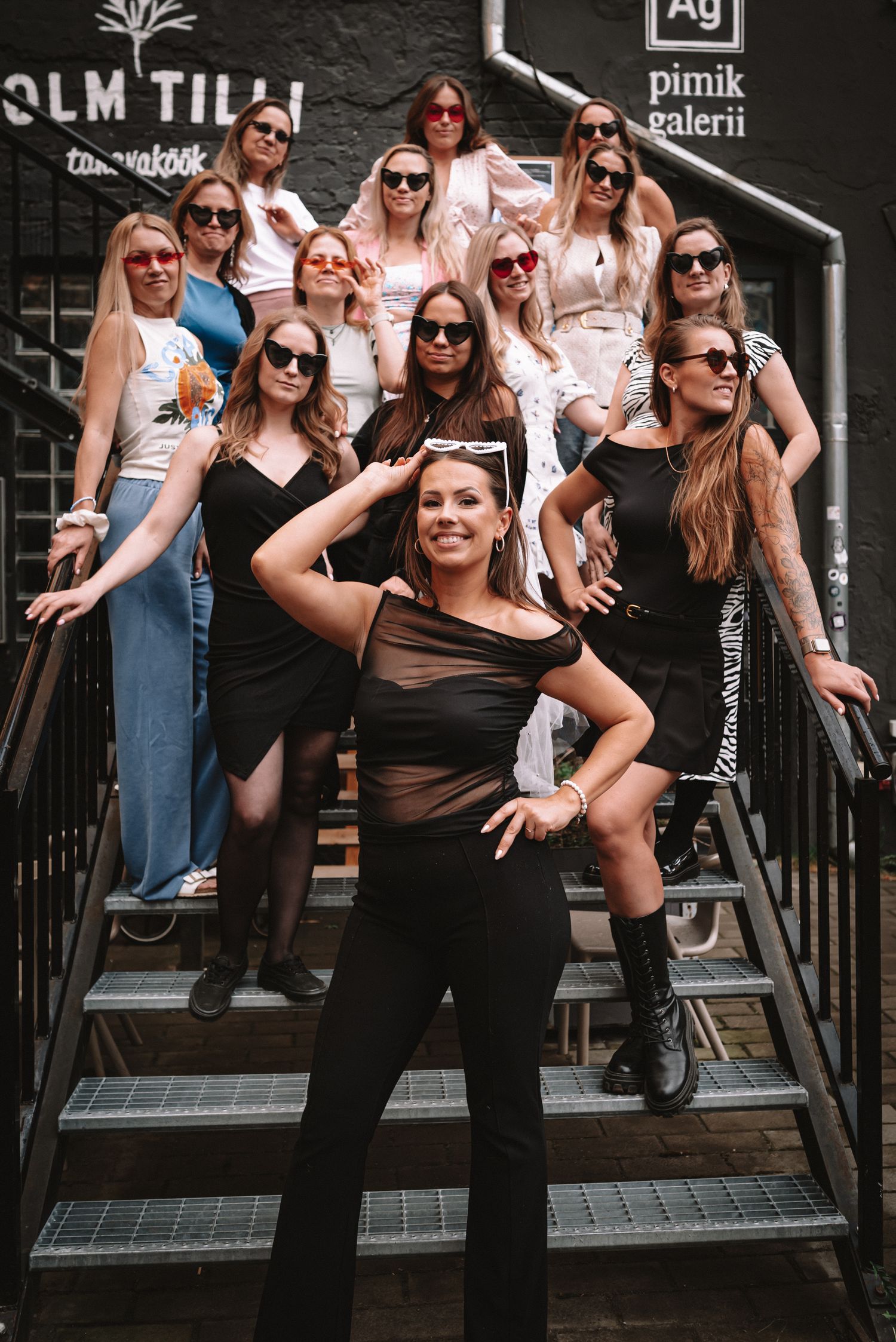 A group strike stylish poses on stairs outside a venue wearing coordinated black and white outfits and sunglasses.