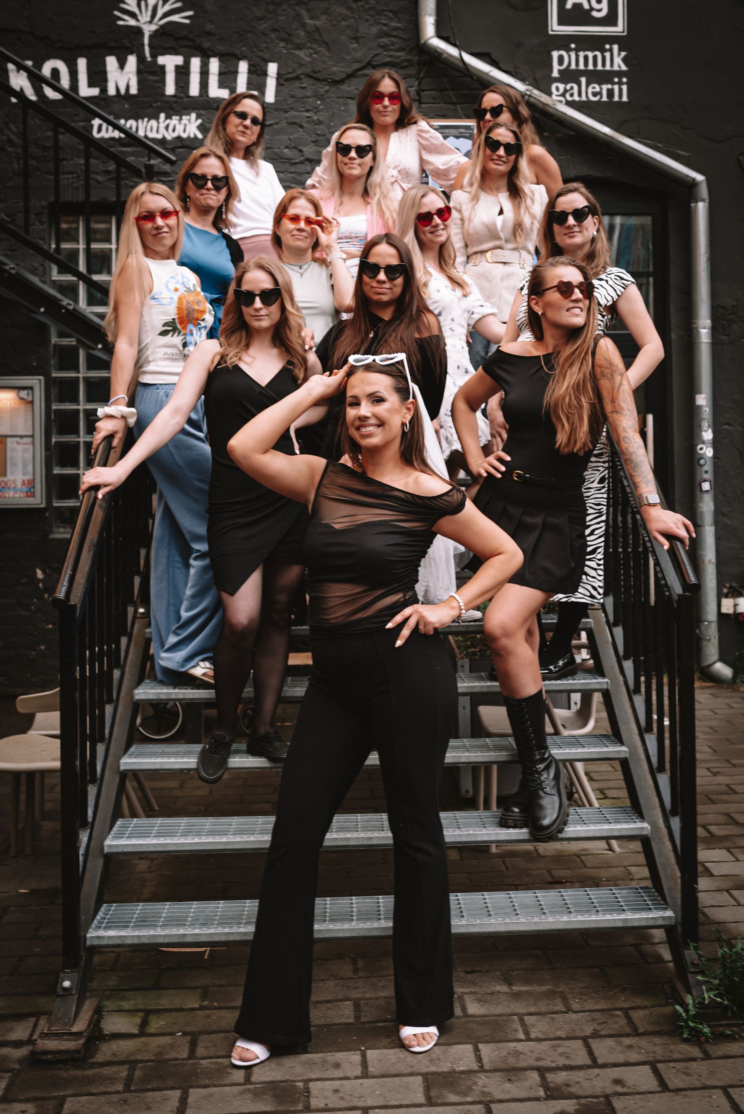 Group poses on outdoor wooden staircase in summer outfits and sunglasses outside brick building.