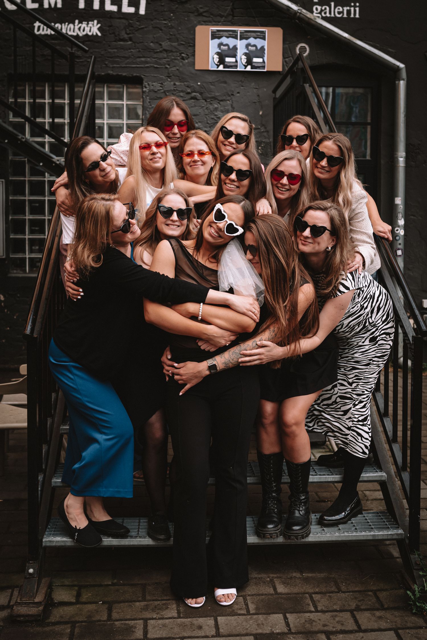 A group of ten friends wearing sunglasses pose together on stone steps outside a dark building entrance.
