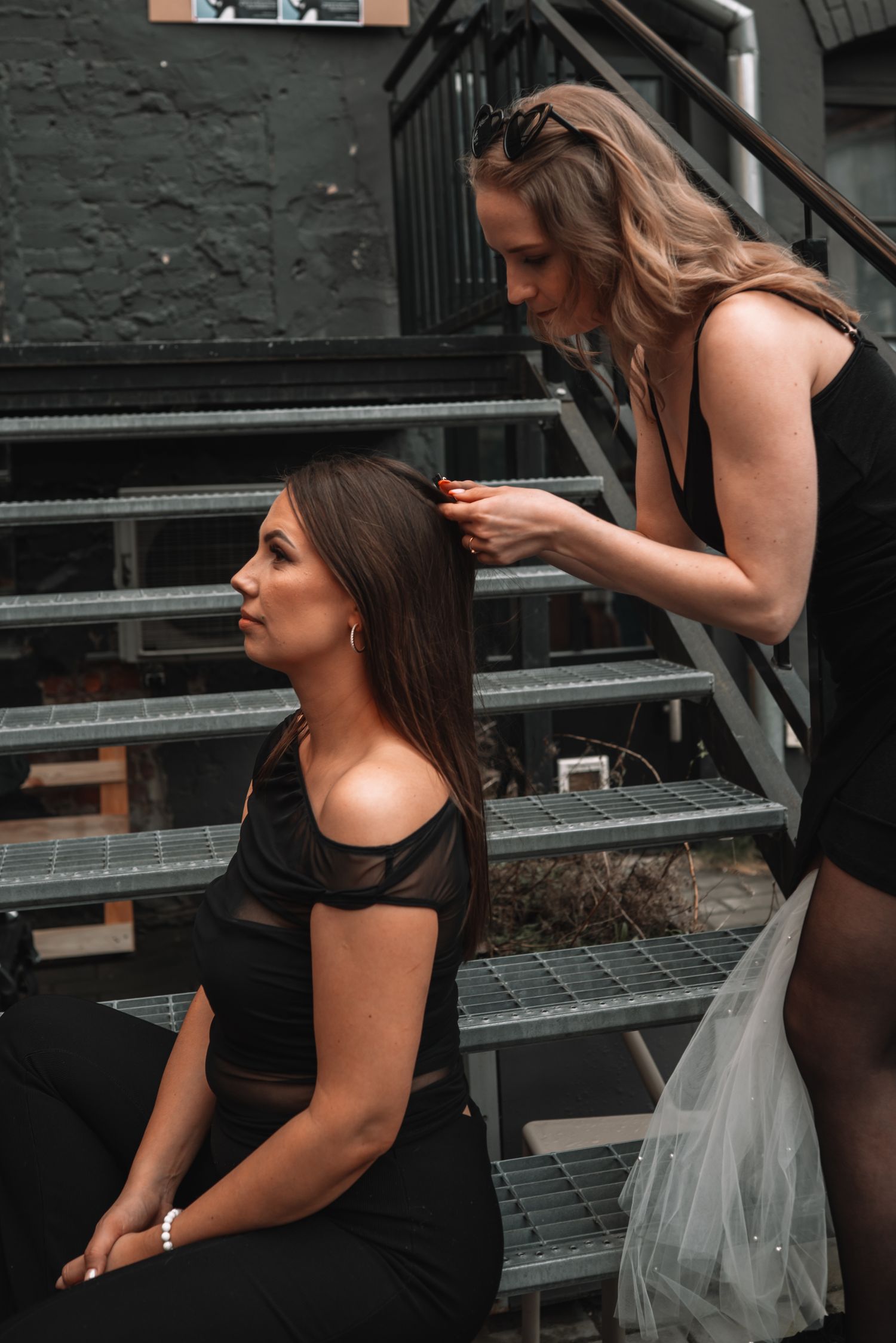 Two people in black outfits sit on industrial metal stairs while styling hair before an event.