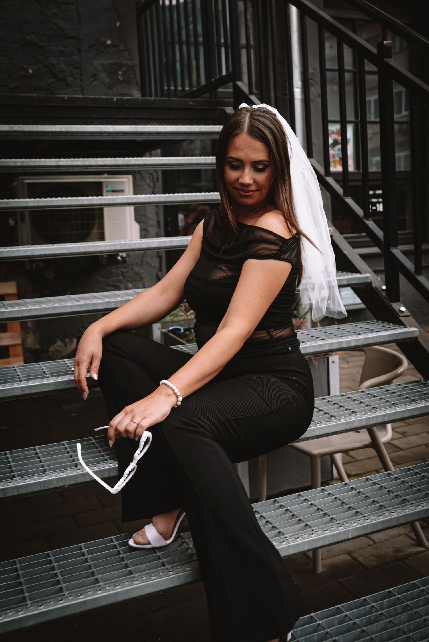 A person in a black outfit and white veil sits on industrial metal stairs holding a phone.