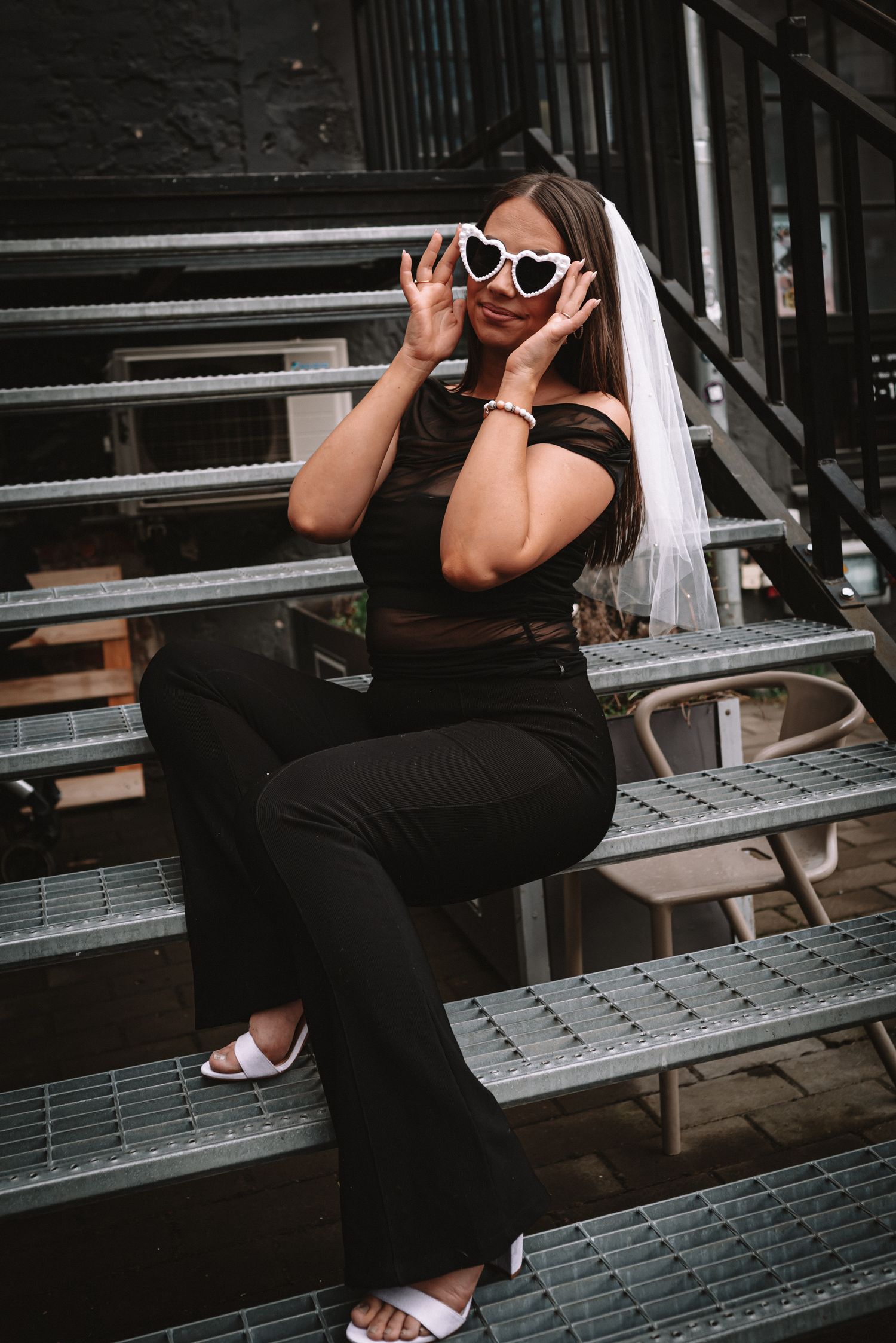 A person in white sunglasses and black attire sits on metal stairs wearing a veil and striking a playful pose.